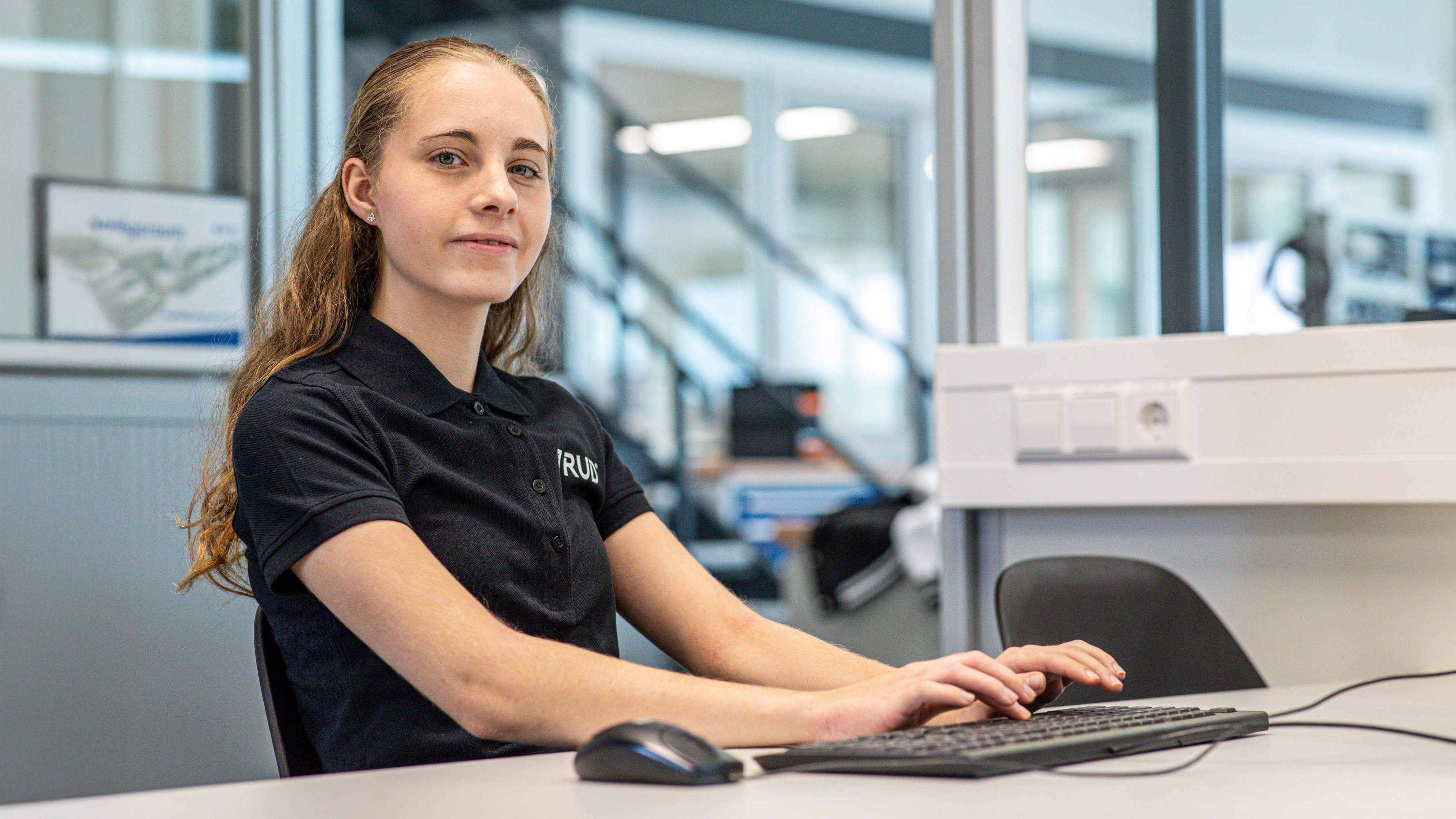 Junge Frau in schwarzem RUD Polohemd sitzt am Schreibtisch und arbeitet an Computer mit Tastatur und Maus in einem modernen Büro.