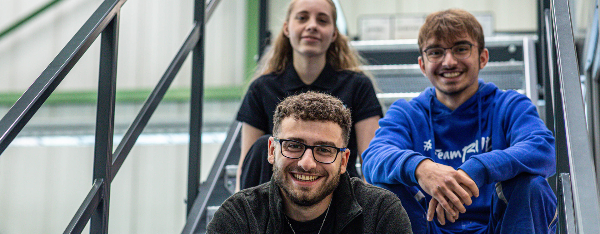 Drei junge RUD Mitarbeitende sitzen lächelnd auf einer Treppe in einem industriellen Arbeitsumfeld.