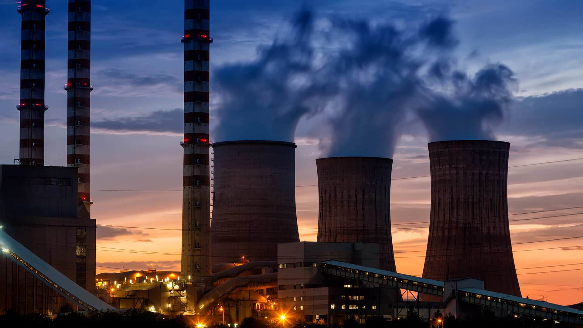 Industriekraftwerk mit Kühltürmen und Schornsteinen bei Sonnenuntergang, Dampf steigt in den Abendhimmel.
