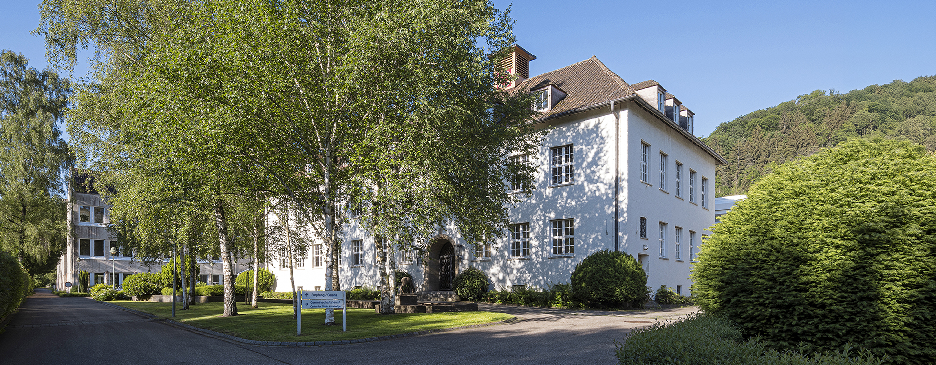 Das weiße RUD Campus-Gebäude mit Satteldach und vielen Fenstern, umgeben von Bäumen und Grünflächen an einem sonnigen Tag.