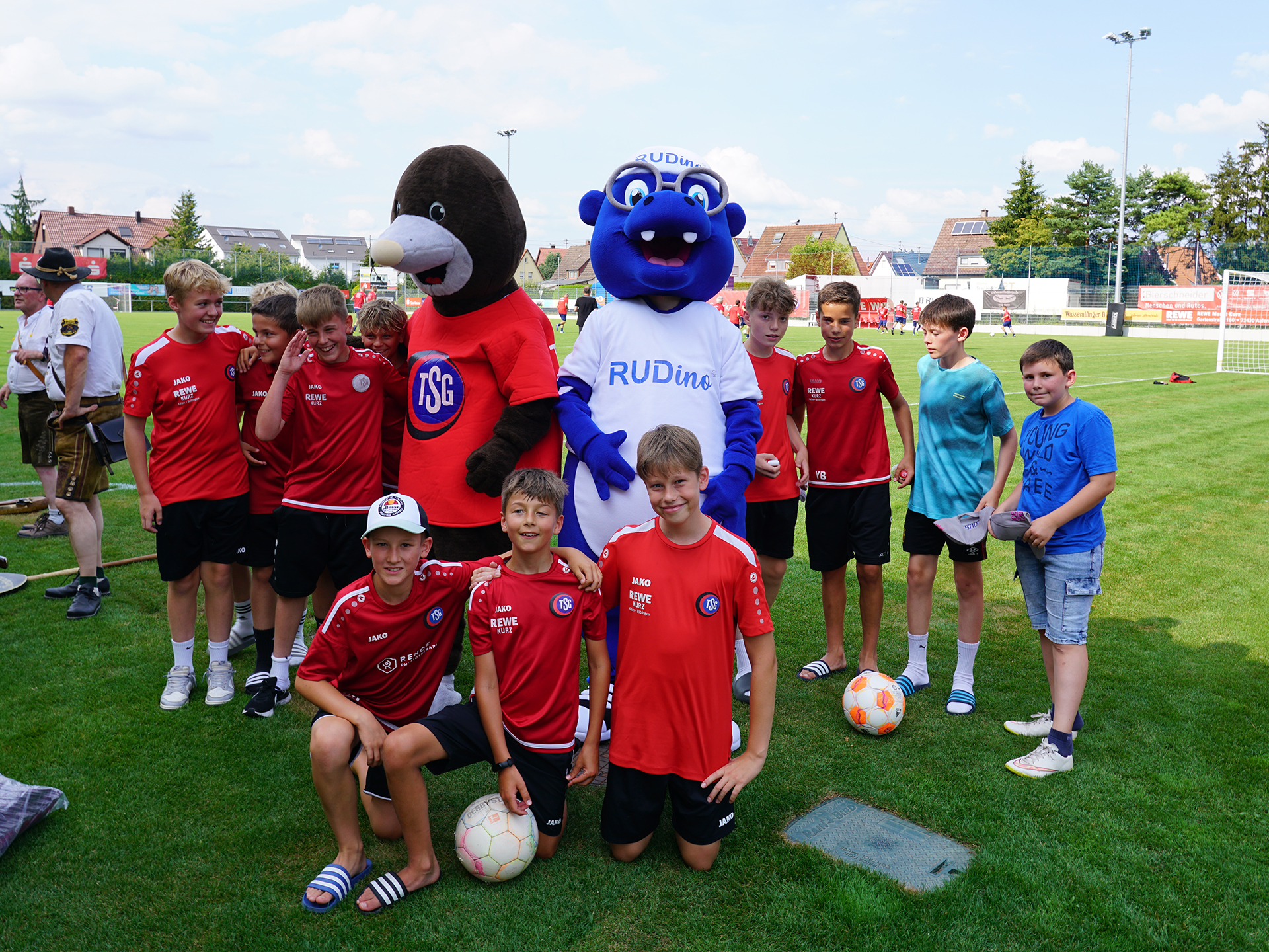 Kinderfußballteam mit den Maskottchen RUDino und TSG-Maulwurf bei einem Sportevent auf dem Fußballplatz.