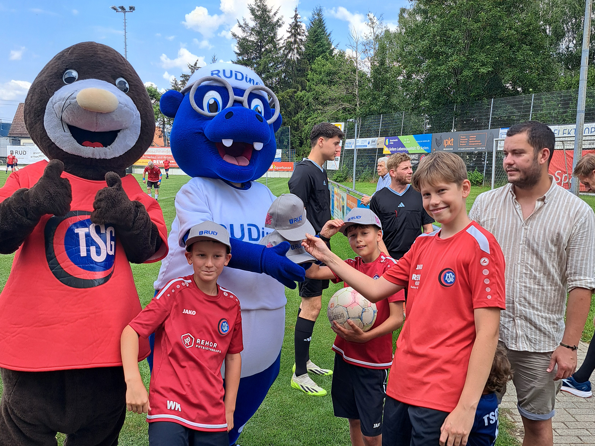 Kinderfußballer mit Maskottchen RUDino und TSG-Maulwurf bei einer Übergabe von RUD-Kappen auf dem Spielfeld.